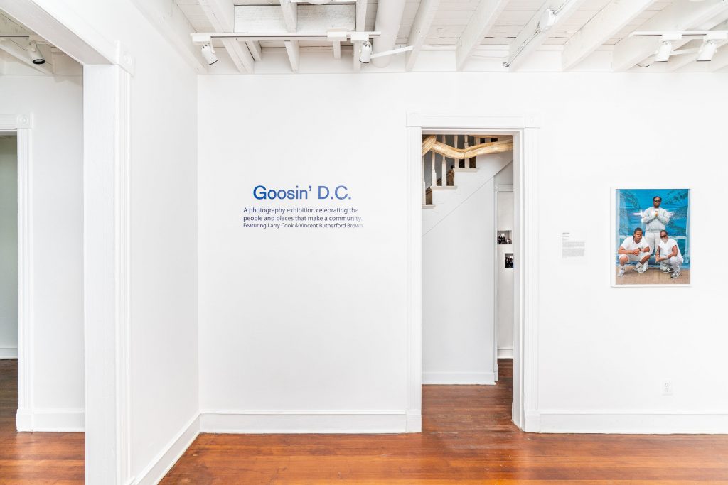 image of a room in The Nicholson Project gallery. original floors, white walls and ceiling are punctuated by an applied exhibit description, "Goosin' DC" with introductory blurb below. To the right a photograph shows a group of Black men in white posing against a blue background. Through a door in the middle, the hallway and other galleries are visible.