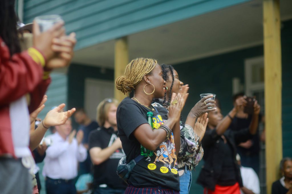 a photograph of The Nicholson Project opening party, outside in the community garden with the teal siding of the house in the background. In the foreground, a diverse group of people clap in varying degrees of focus.